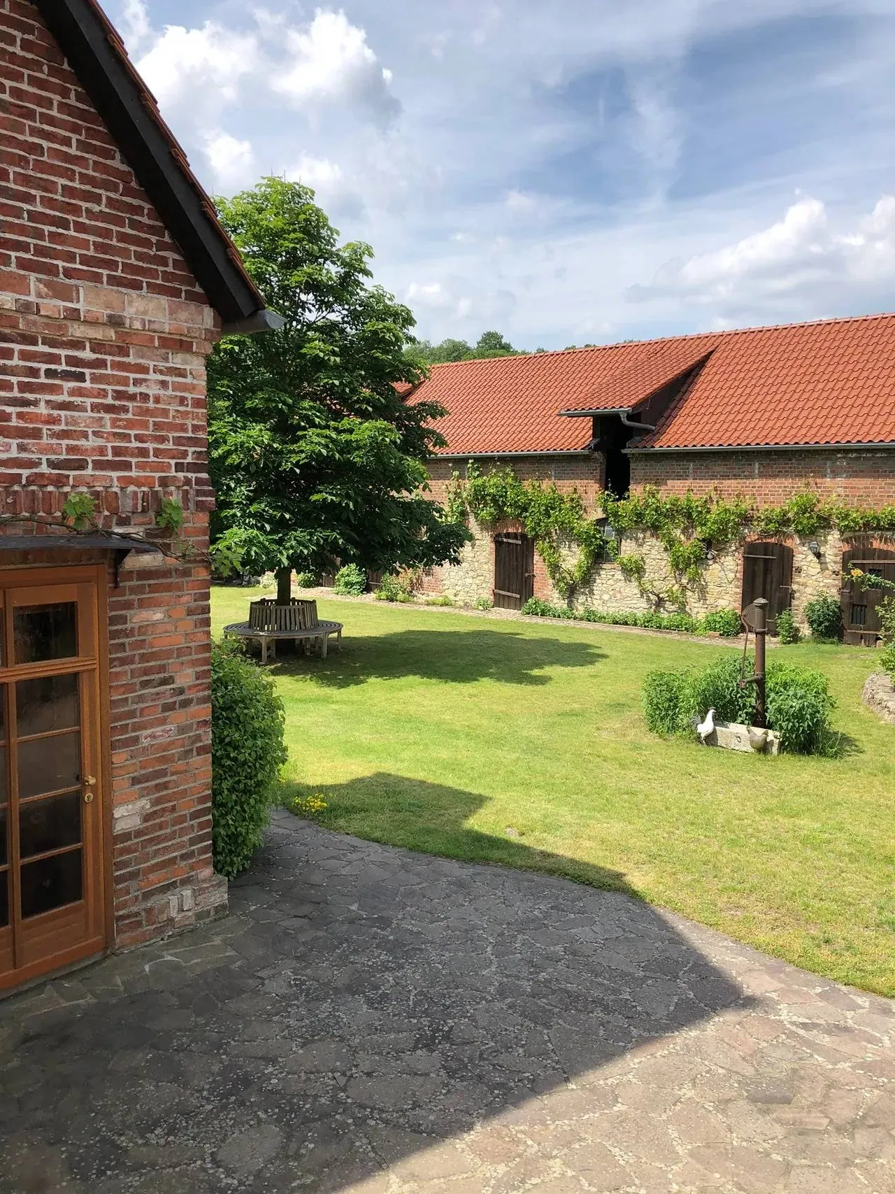 Innenhof des Ferienhofs mit Rasen, Rundbank unter dem Baum und berankter Backsteinscheune
